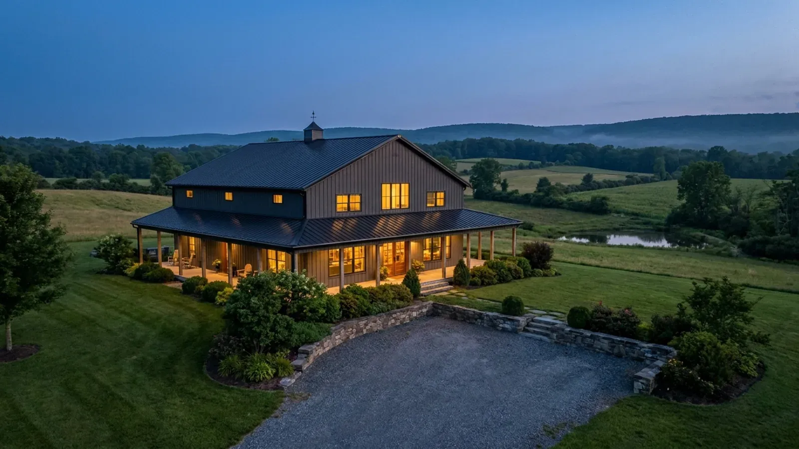 A finished barndominium at dusk with warm interior lighting glowing through its large windows.