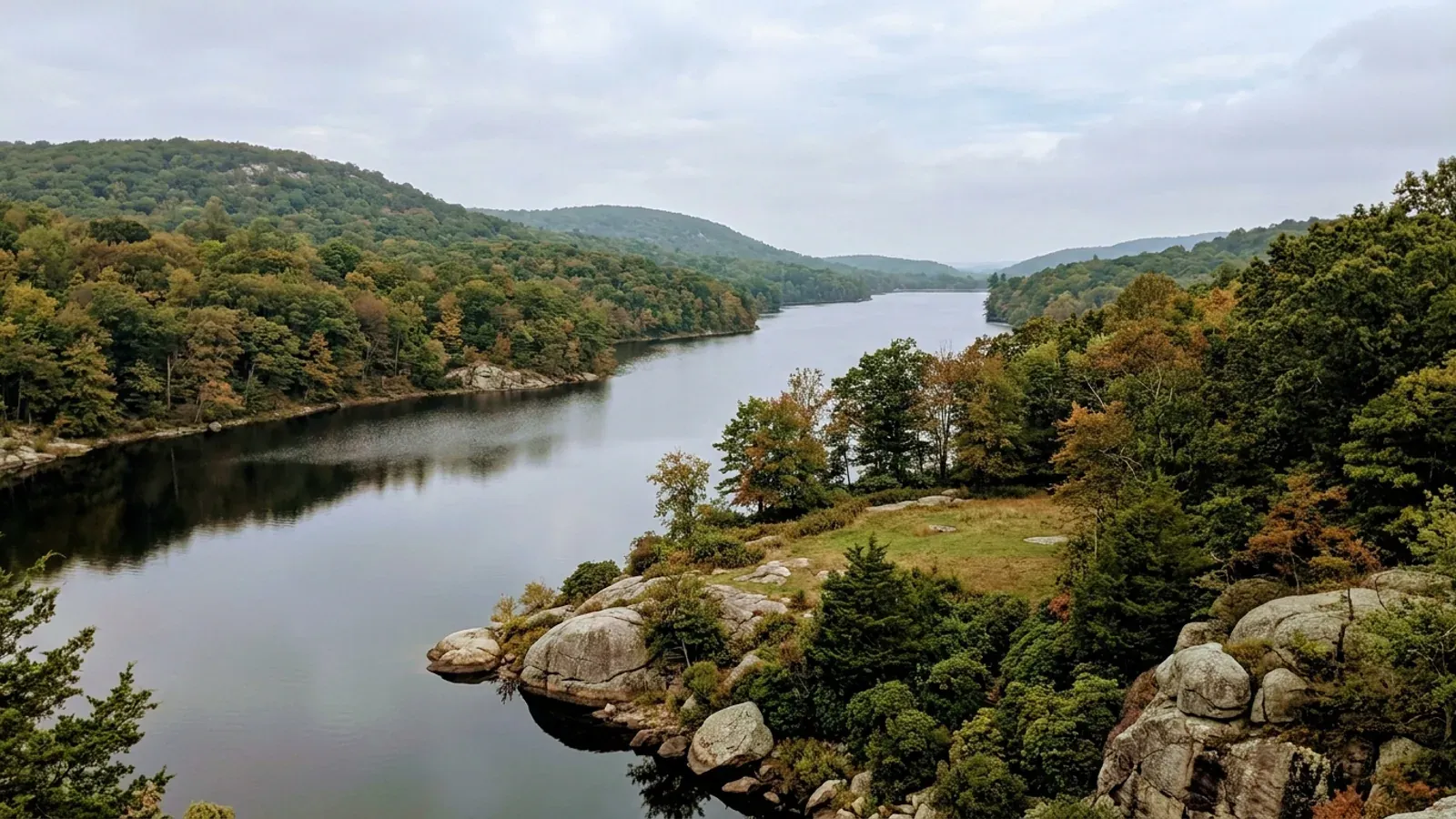 A calm reservoir winding between low forested hardwood hills, with grey boulders and mountain laurel along the rocky shore.