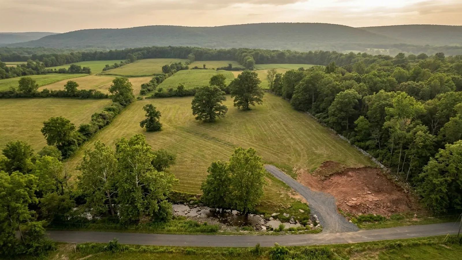 Aerial view of an undeveloped rural lot in a mown hay field, with a gravel drive, a hardwood woodlot and stone wall along one edge, and a forested ridge on the horizon.